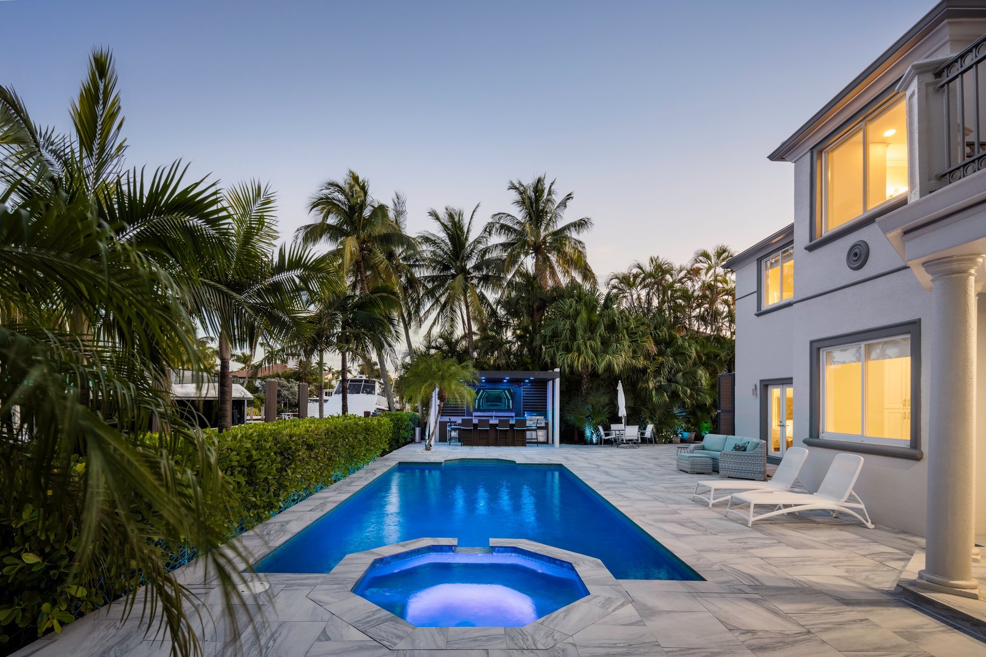Modern colonial-style house with illuminated swimming pool, palm trees, lounge chairs, and outdoor furniture in a luxury residential neighborhood